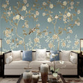 Papier peint panoramique fleuri bleu avec branches dorées et oiseaux derrière un canapé crème